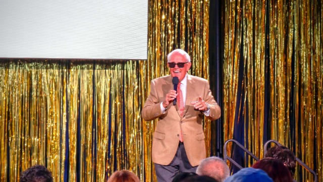 Man in a tan suit and sunglasses speaking on stage with gold curtain backdrop at a conference.