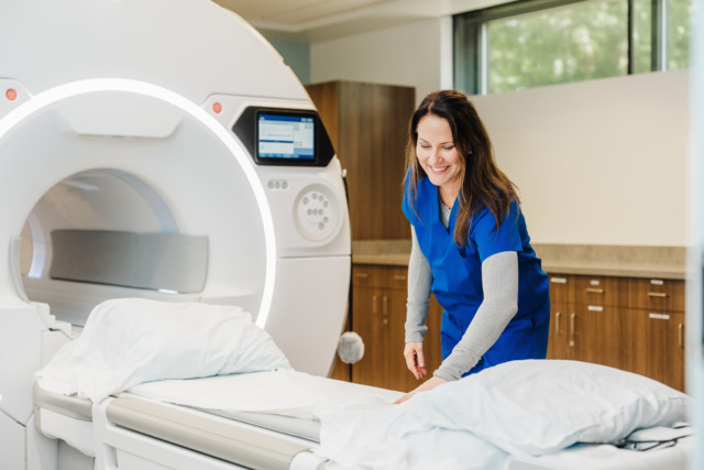 Healthcare professional in blue scrubs smiling beside an MRI machine in a modern medical facility.