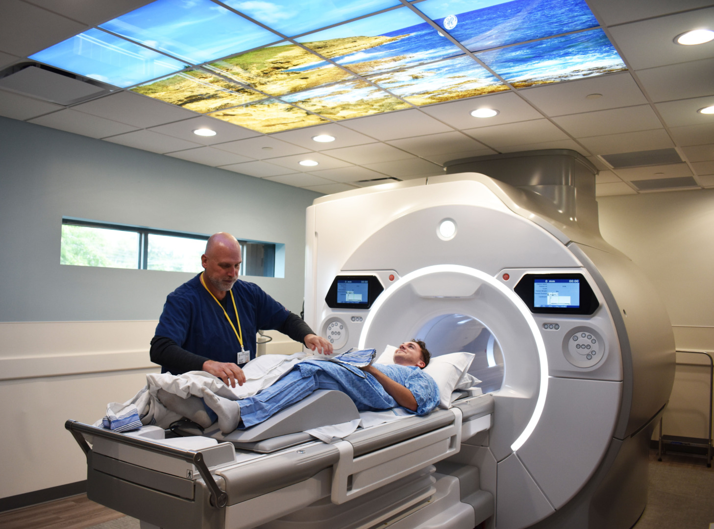 Technician assists patient lying down for an MRI scan with a scenic ceiling display in a modern medical facility.