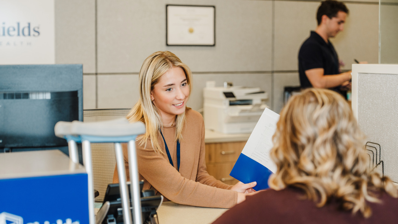 Woman assisting a customer at an office desk with a folder, while another person works in the background.