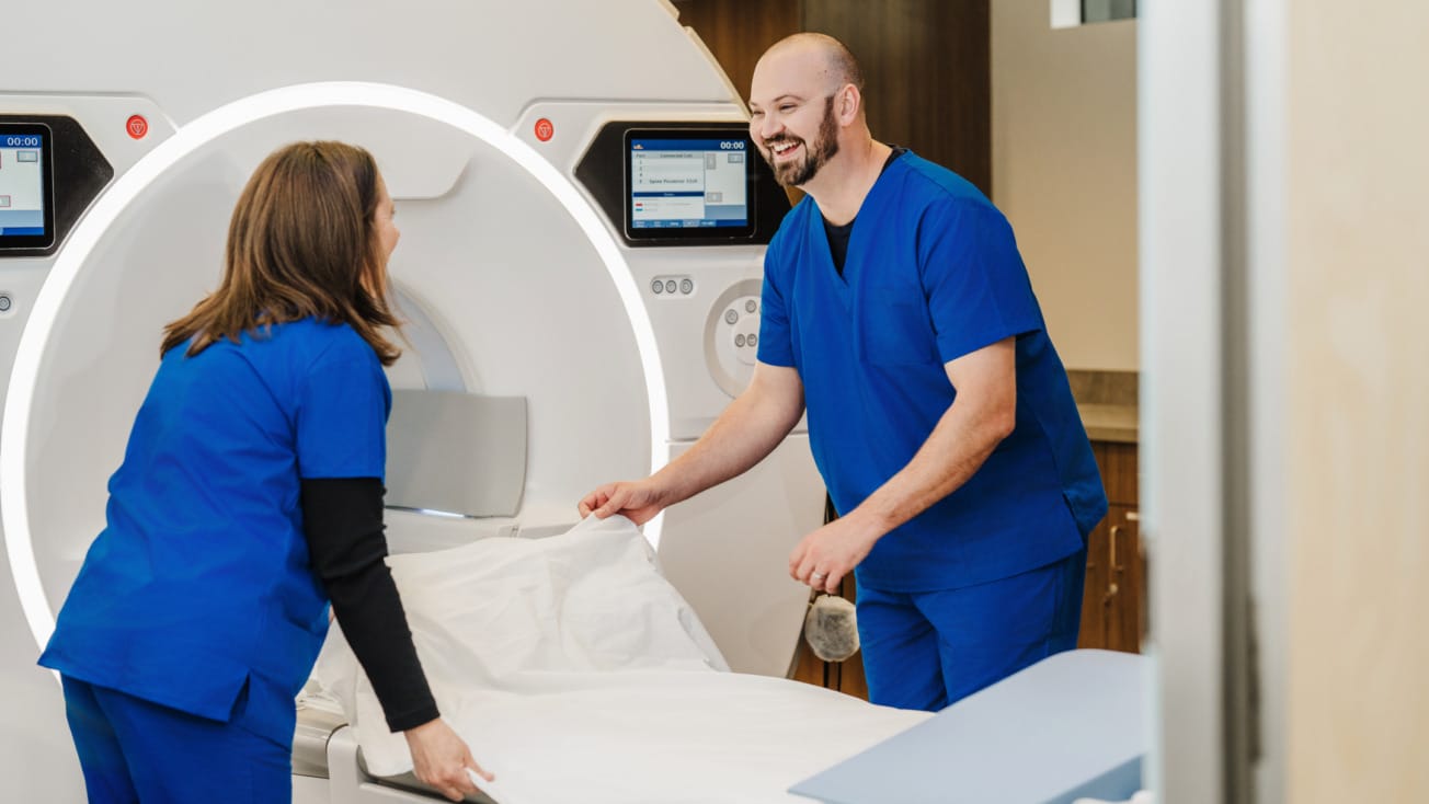 Male and female nurses in blue scrubs smiling as they place a fresh bed liner on a patient table in front of an MRI machine.