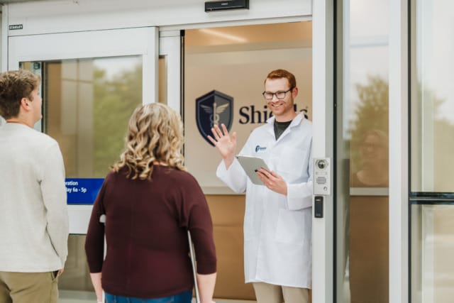 Doctor in a white coat smiling and greeting two people at a clinic entrance while holding a tablet.