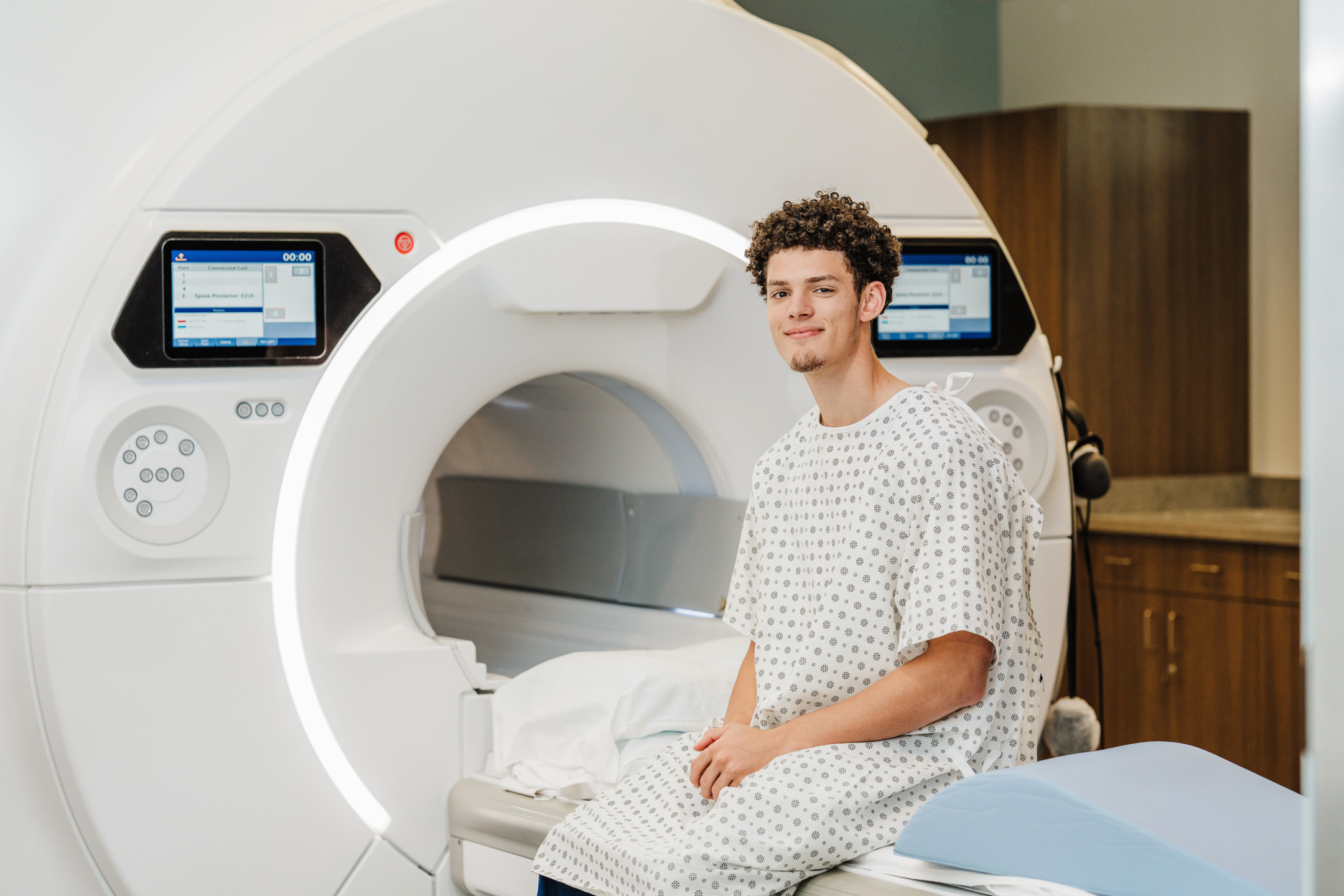 Person in a hospital gown sitting on an MRI machine inside a medical facility.