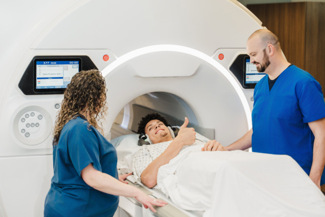A patient lying on a hospital bed gives a thumbs up while about to enter an MRI machine, with two nurses in blue scrubs standing on each side supportively.