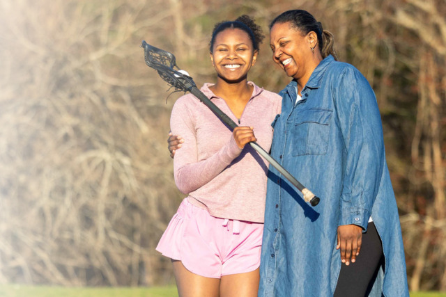 Mother and daughter smiling outdoors, daughter holding a lacrosse stick, both enjoying a sunny day.