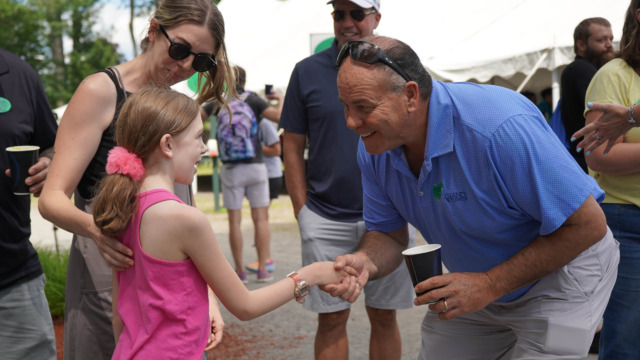 Man in blue shirt shaking hands with a young girl at an outdoor event, surrounded by people and tents in the background.
