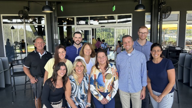 Group of people smiling and posing together inside a modern room with large windows and visible outdoor scenery.