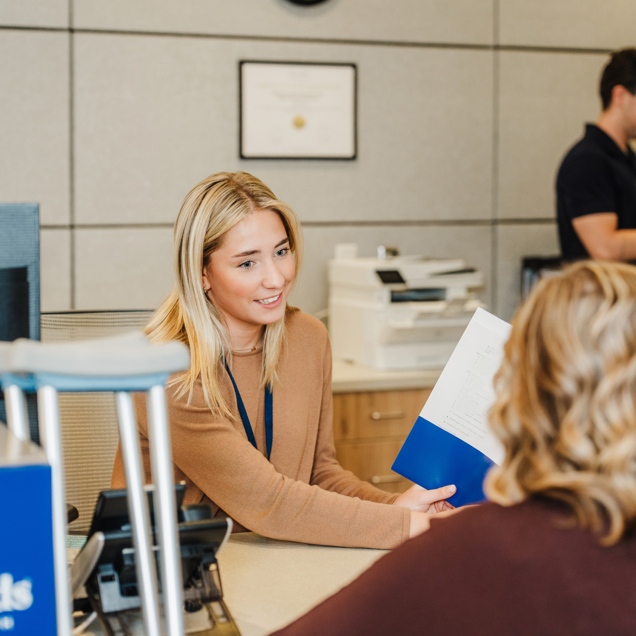Receptionist assisting a customer at a service desk with paperwork, while a man works in the background on a computer.