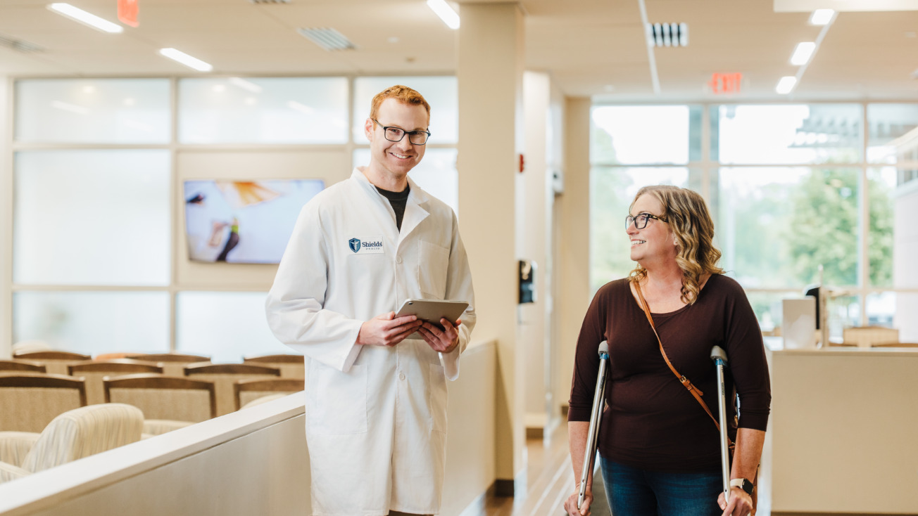 Doctor in a white coat smiles while talking to a woman with crutches in a bright, modern medical office.