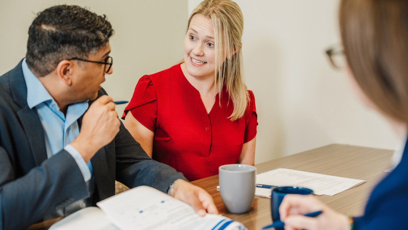 Three professionals having a discussion at a table with documents, one person in a red shirt actively speaking.