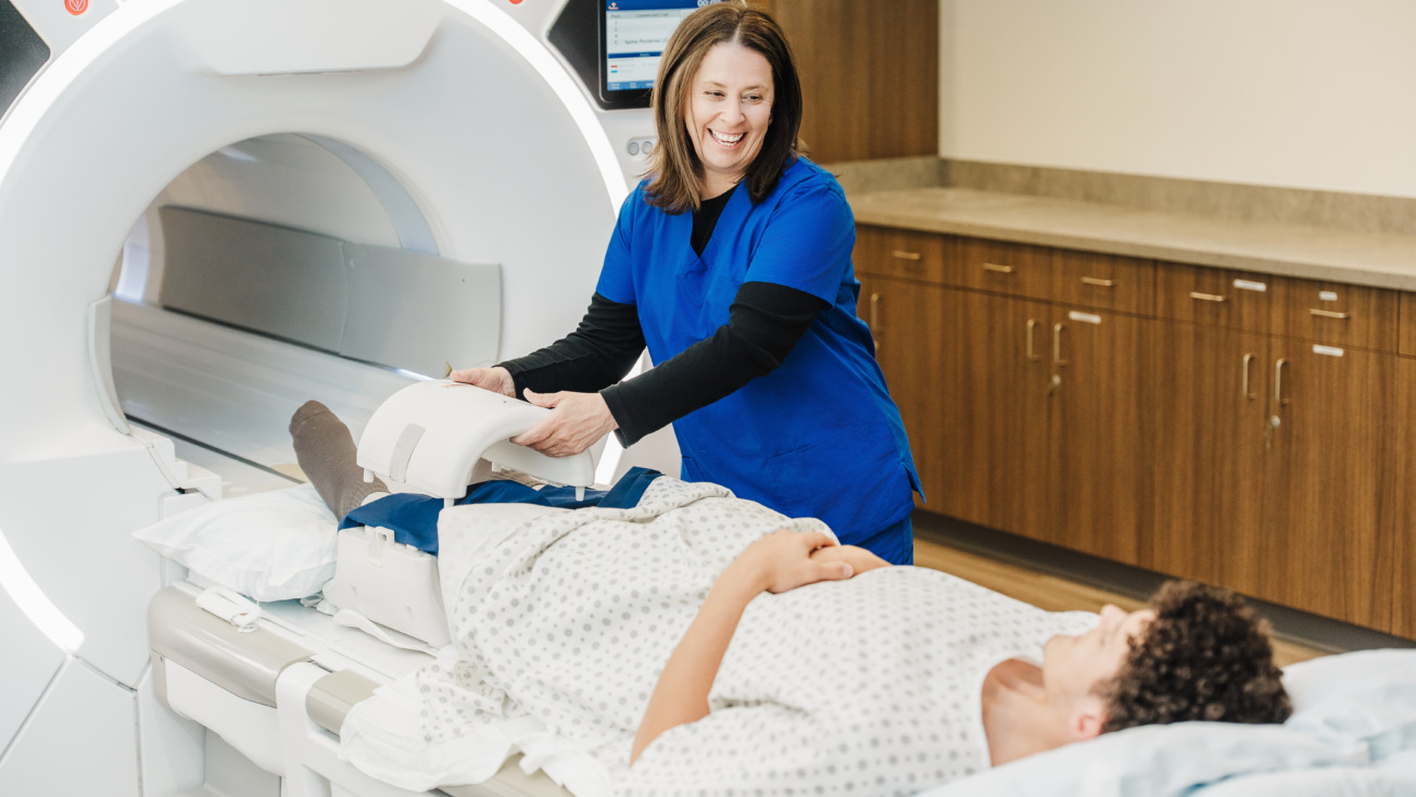Smiling female nurse in blue scrubs assisting a patient in a hospital gown as they prepare to enter an MRI machine.