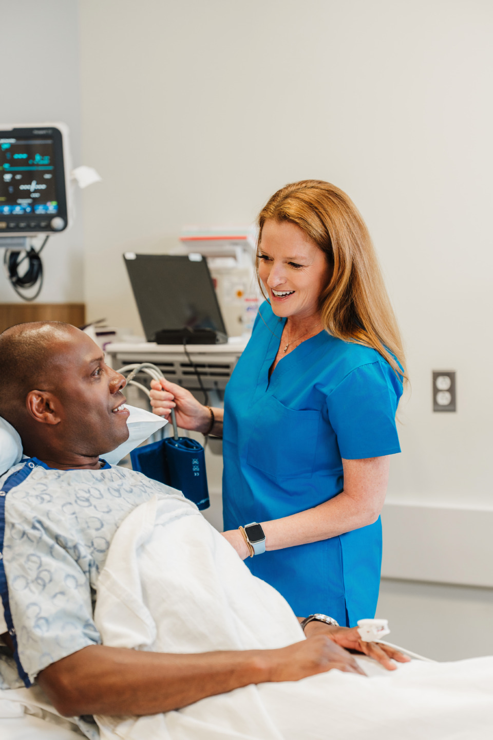 Nurse in blue scrubs speaking with a patient lying in a hospital bed, medical charts and equipment in the background.