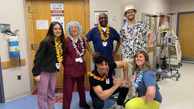Hospital staff in colorful leis pose together, smiling in a hallway decorated with medical equipment and motivational posters.