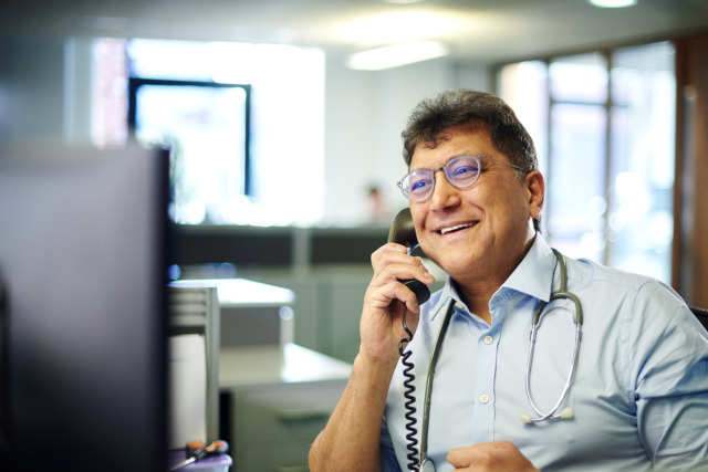 Smiling professional in an office with telephone and stethoscope around neck, looking at a computer monitor.
