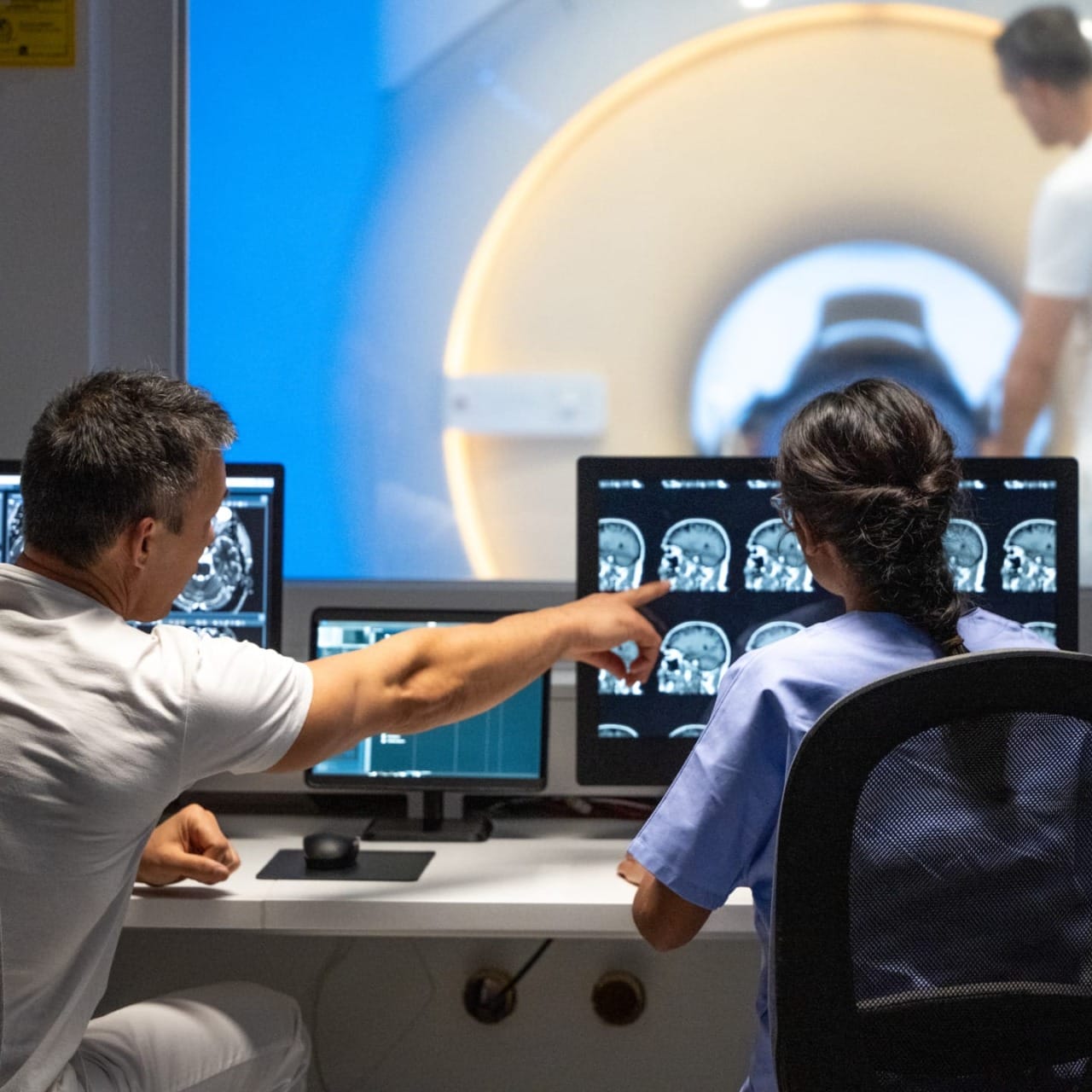 Two medical professionals analyzing brain scans on computer screens in a hospital radiology room with an MRI scanner in view.