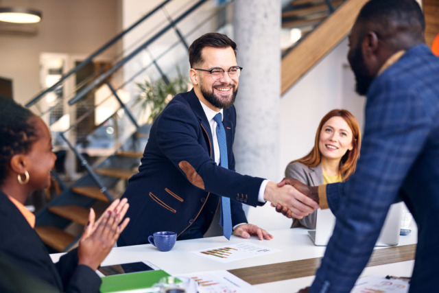 Business professionals shaking hands in office meeting, diverse group, charts on table, stairs in background.