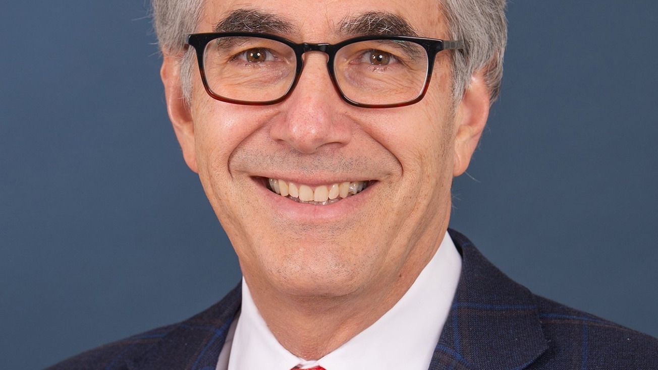 Man wearing glasses, a blue suit, white shirt, and red tie, smiling with a blue background.