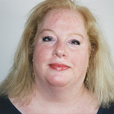 Person with light hair and fair skin, wearing a dark top, smiling slightly, and looking at the camera against a plain background.