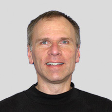 Smiling person with short hair wearing a black shirt against a plain background.