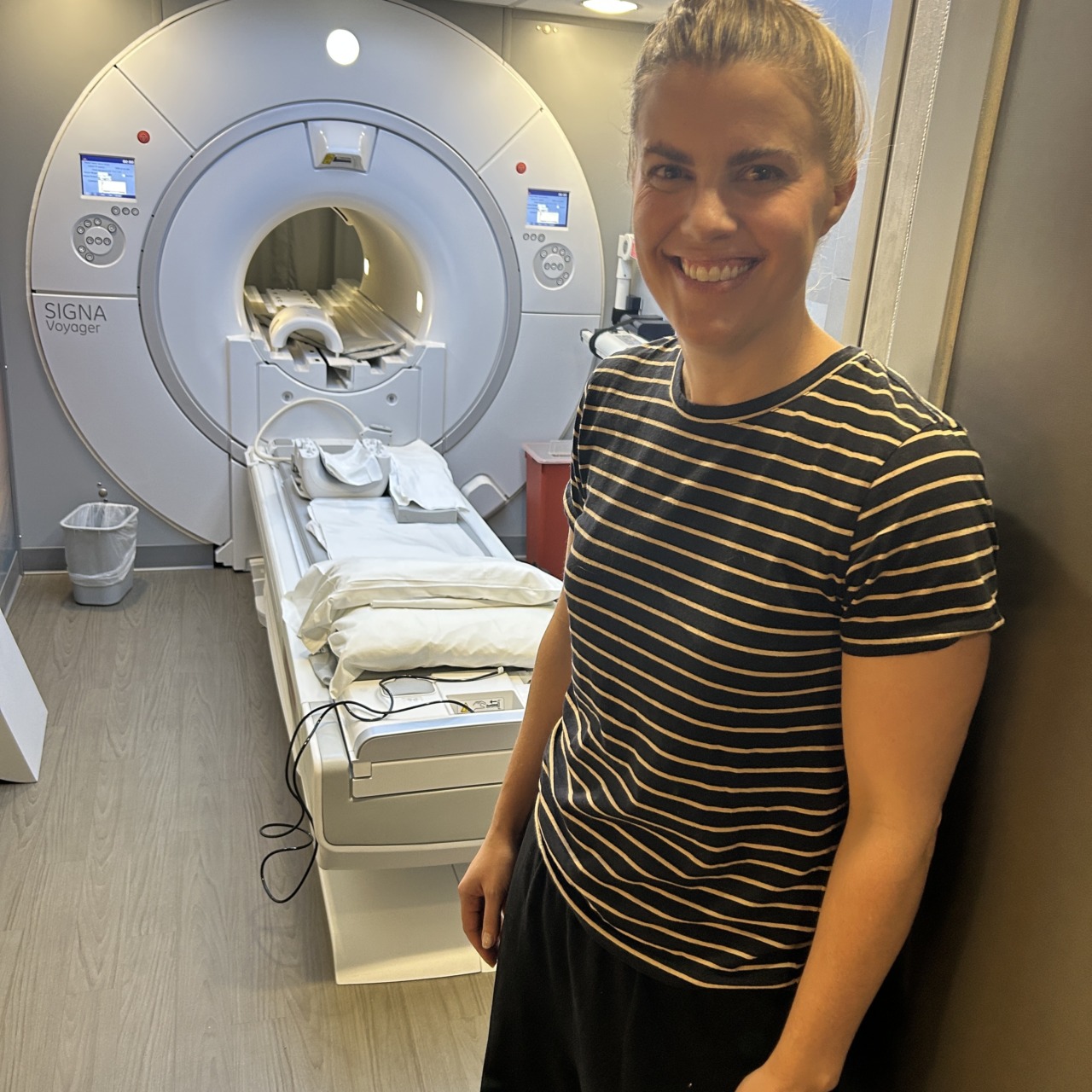 Person standing next to a medical imaging machine in a clinical setting, wearing a striped shirt and smiling at the camera.