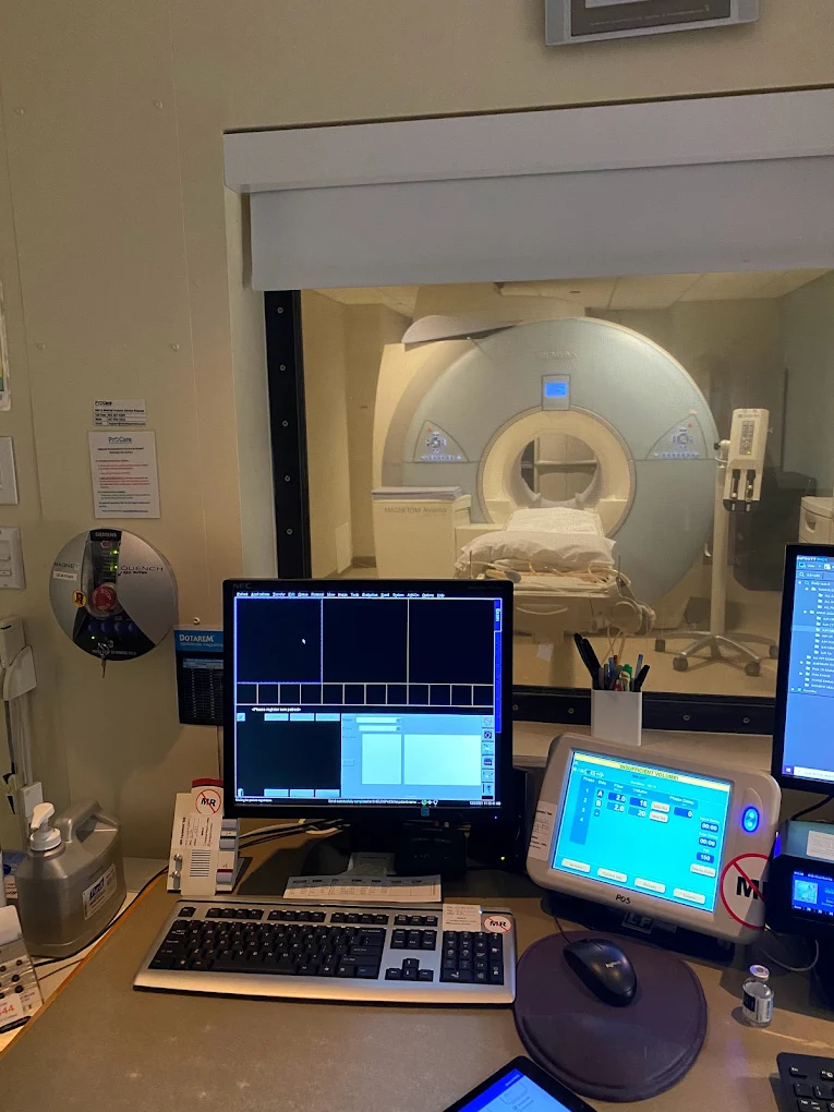 Hospital MRI room with monitors and control panel in the foreground, MRI machine visible through the window in the background.