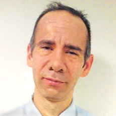 Portrait of a man with short hair and a neutral expression, wearing a light-colored shirt against a plain background.