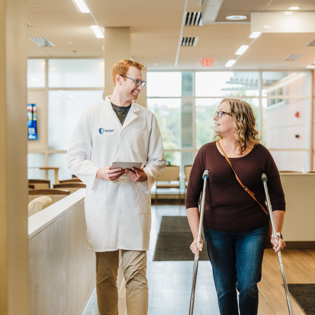 Doctor and patient on crutches walking in a clinic hallway, both smiling and engaged in conversation.