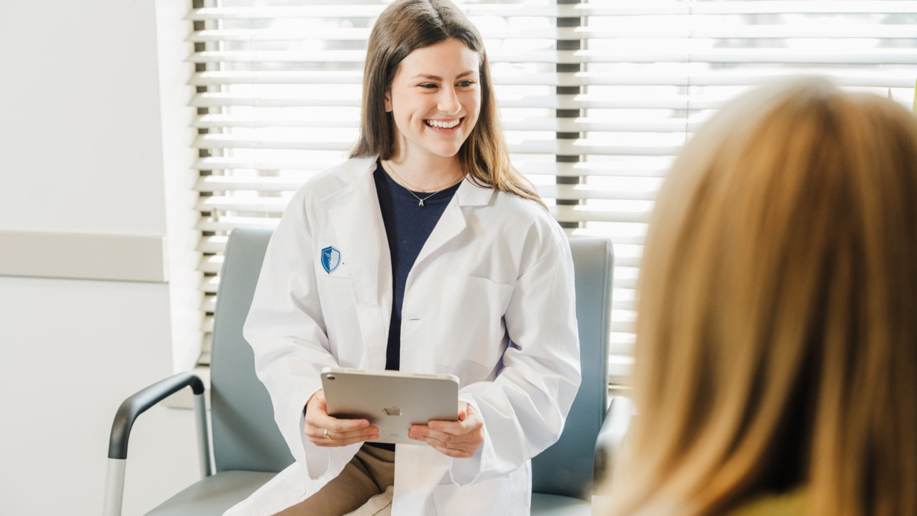 Doctor in a white coat smiling while holding a tablet and talking to a patient in a bright office setting.