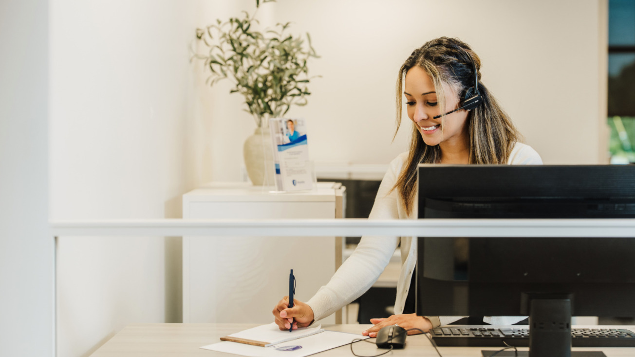 Receptionist with headset writing on a notepad at a desk with a computer and potted plant nearby.