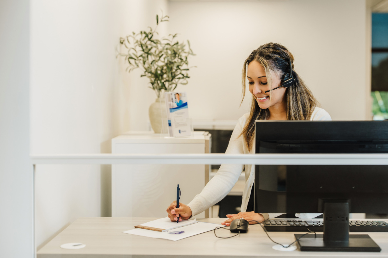 Receptionist with headset writing on a notepad at a desk with a computer and potted plant nearby.