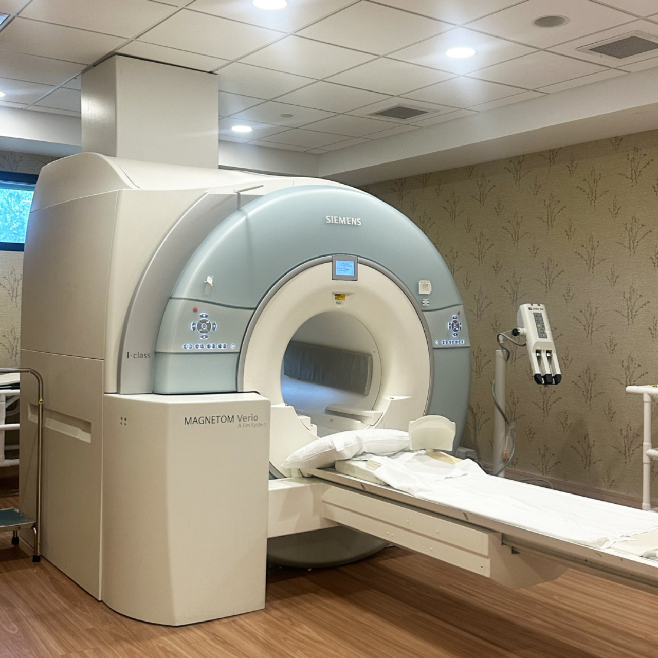 MRI machine in a medical room with wood flooring and patterned wallpaper, ready for patient use.
