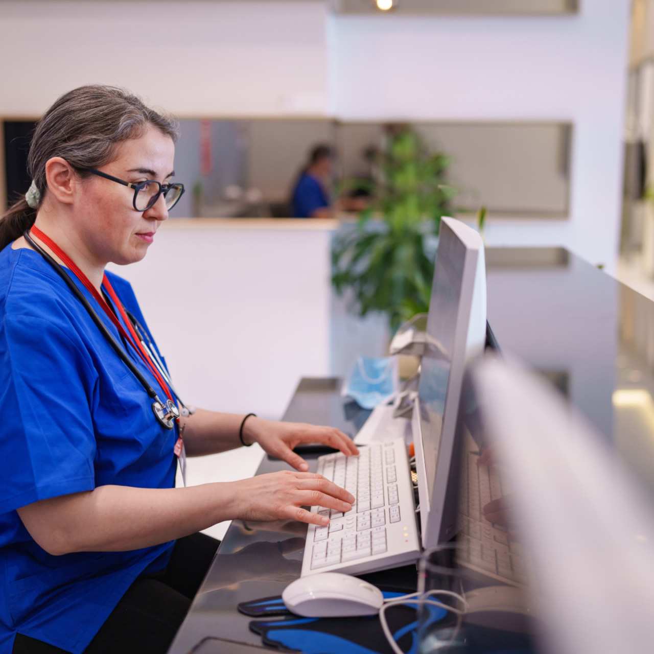 Nurse in blue scrubs working at a computer desk in a medical office setting with plants and modern decor in the background.