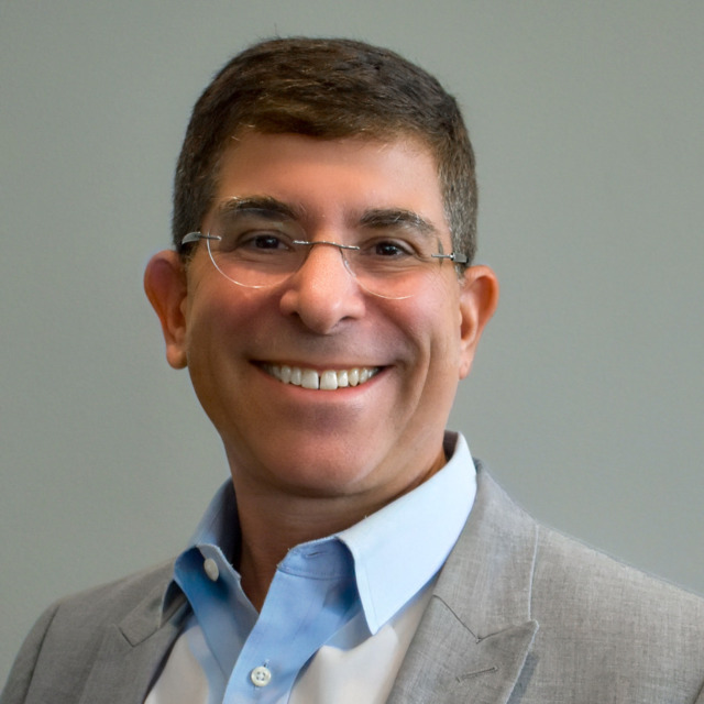 Smiling person wearing glasses and a grey suit jacket against a plain background.