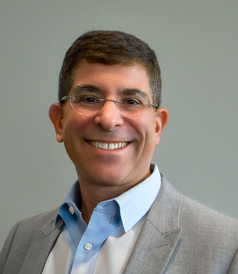 Smiling person wearing glasses and a grey suit jacket against a plain background.