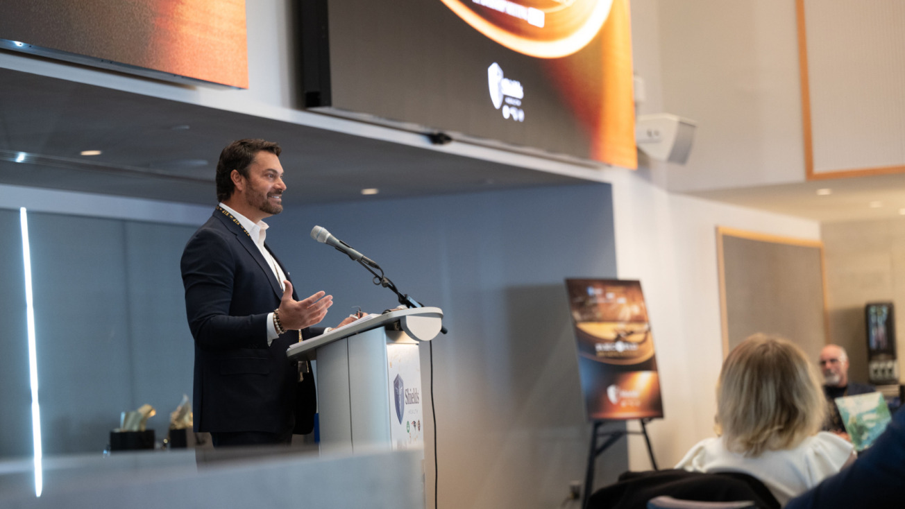 Man speaking at a podium during a conference with presentation screen visible in the background.