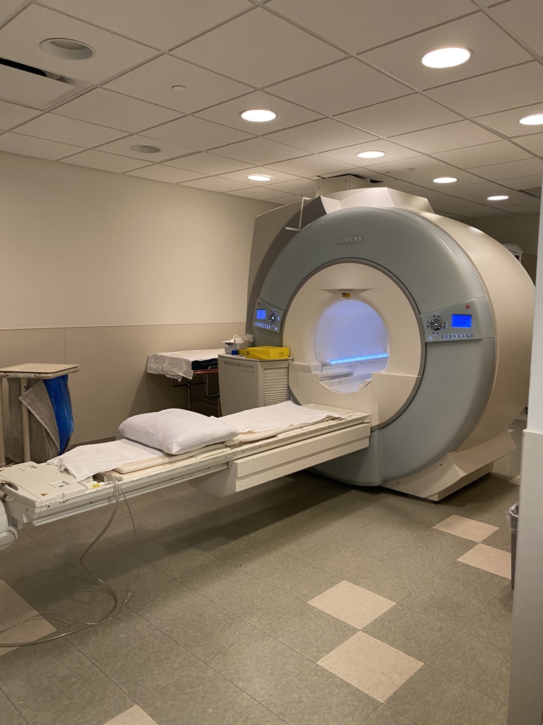 MRI machine in a hospital room with an empty patient bed and medical equipment nearby.