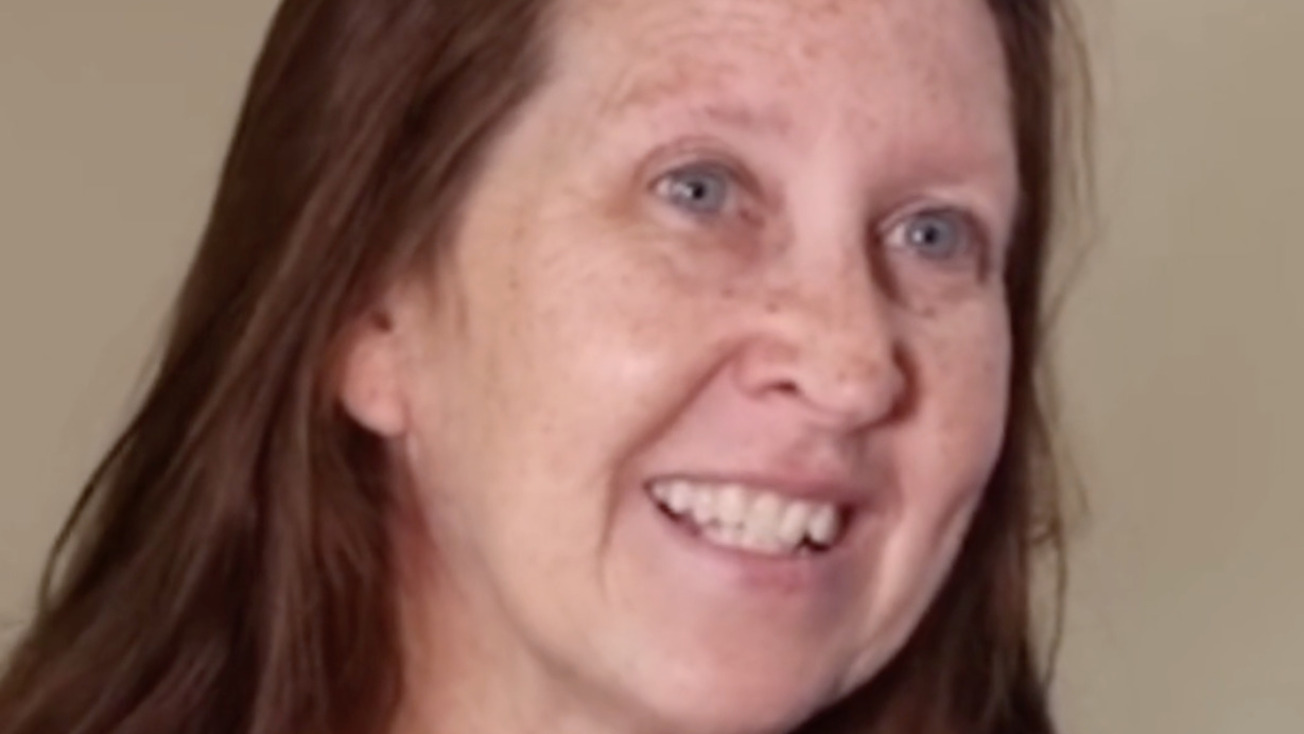 Smiling person with long brown hair wearing a blue top in a light-colored room