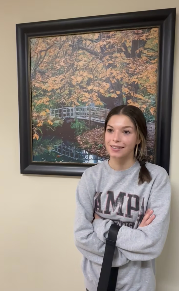 Person standing indoors in front of framed autumn landscape picture, wearing a sweatshirt and smiling with arms crossed.