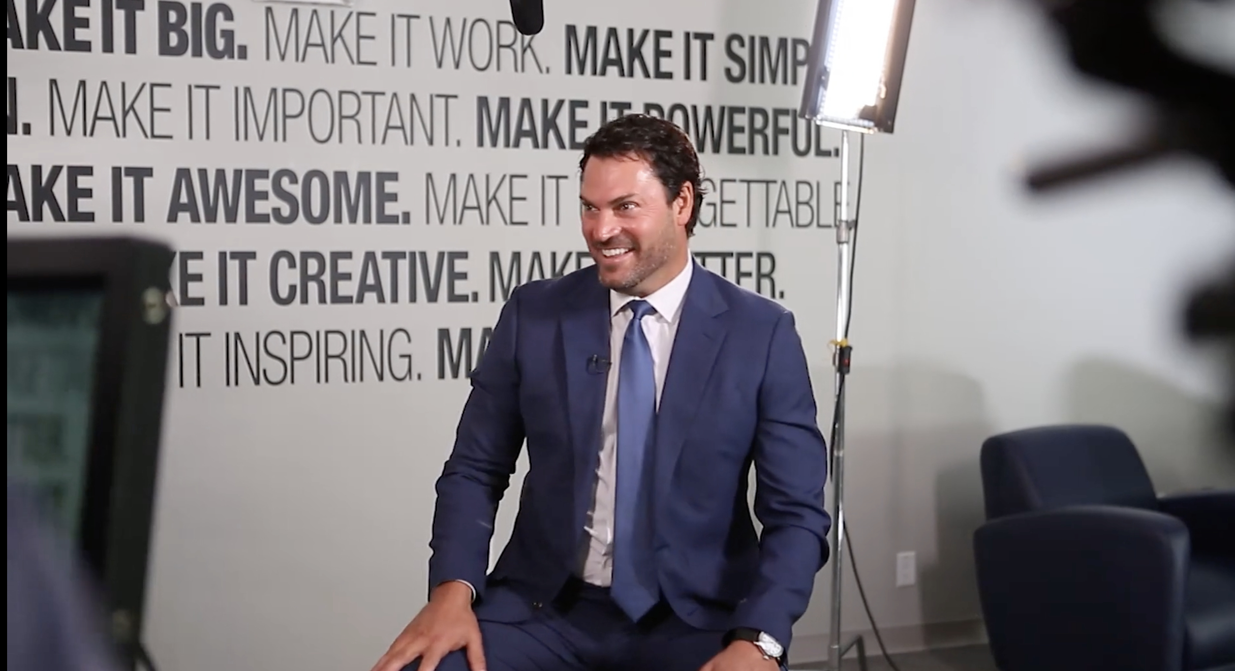 Man in a blue suit sitting and smiling, with motivational words on the wall behind him and a studio light nearby.