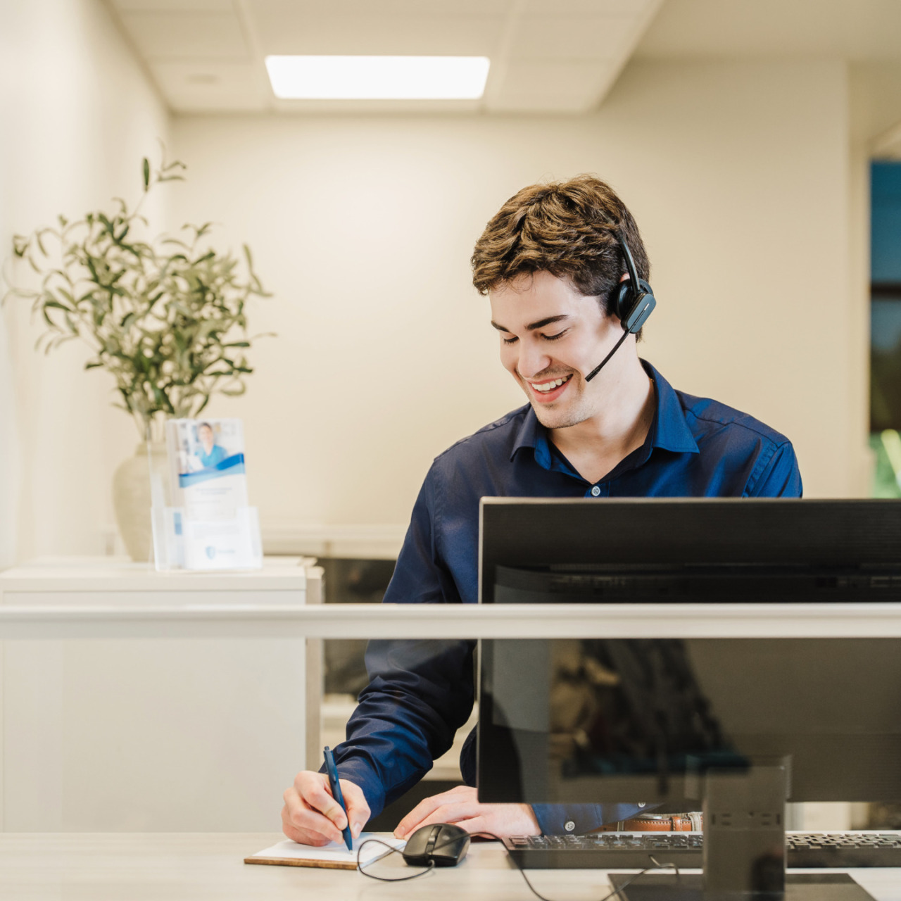 Man with a headset smiling at a desk while writing notes by a computer in a modern office environment.