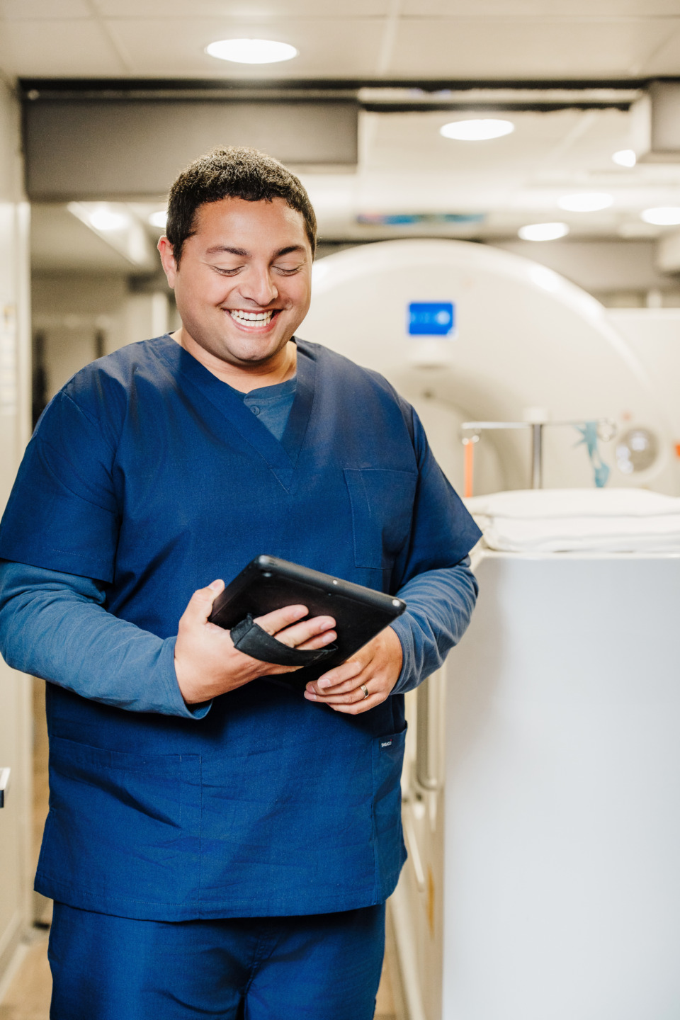 Smiling healthcare professional in blue scrubs holding a tablet in a medical facility.