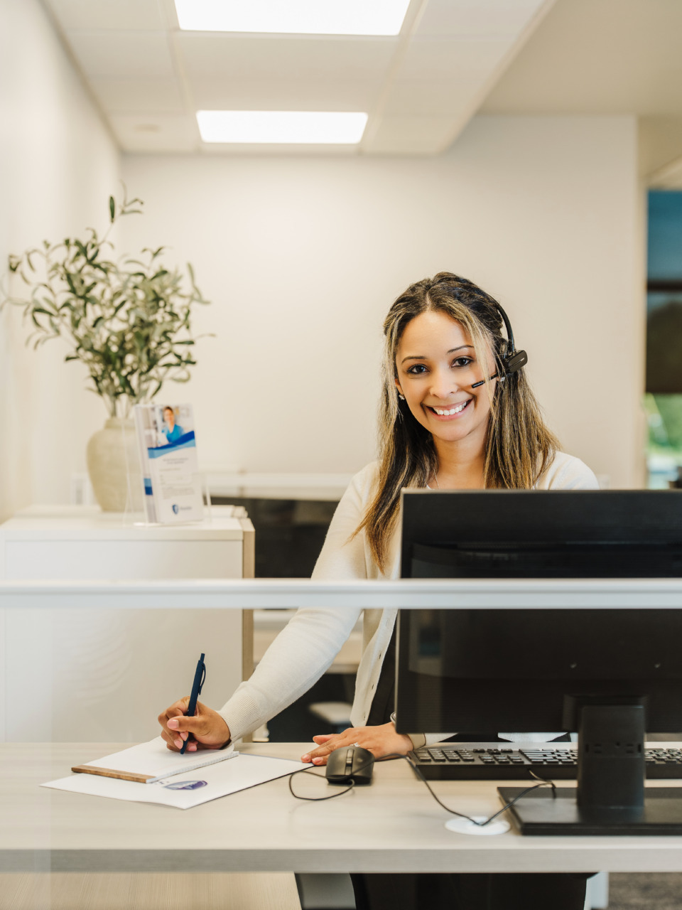 Smiling customer service representative wearing a headset while working at a desk with a computer and notepad.
