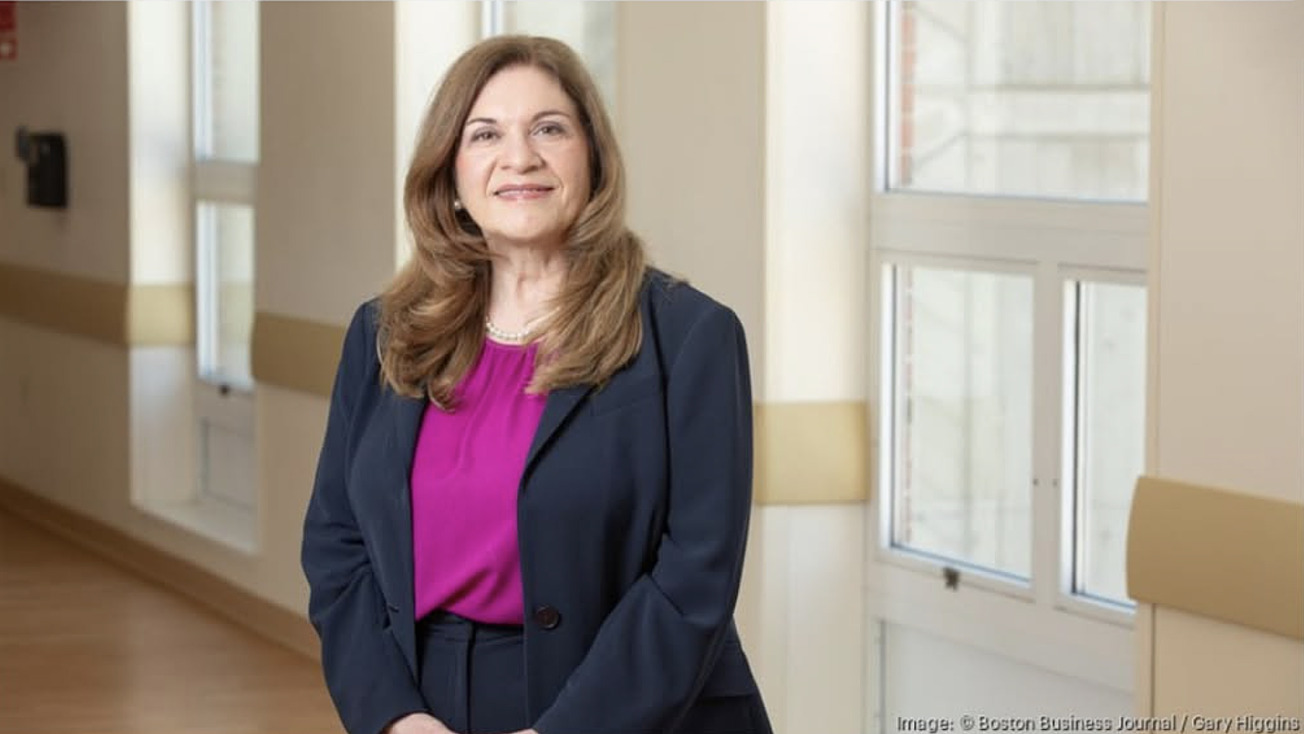 Smiling woman in business attire with long brown hair standing in a well-lit office hallway near large windows.