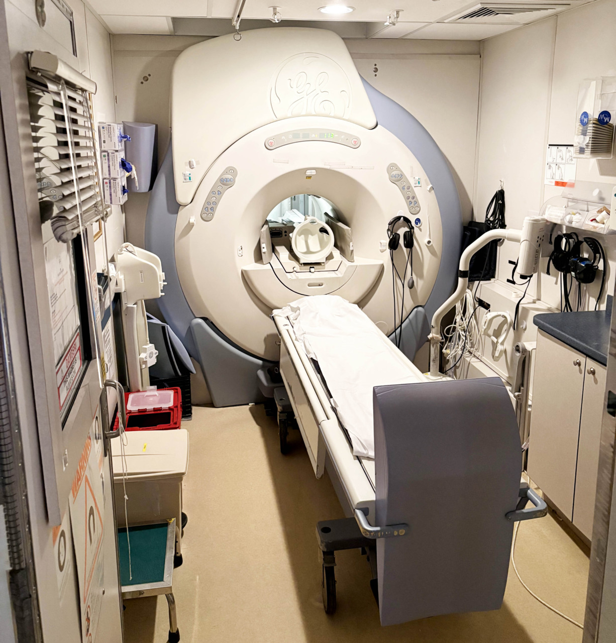 Medical imaging room with an MRI machine surrounded by various medical equipment and a padded patient table in the center.