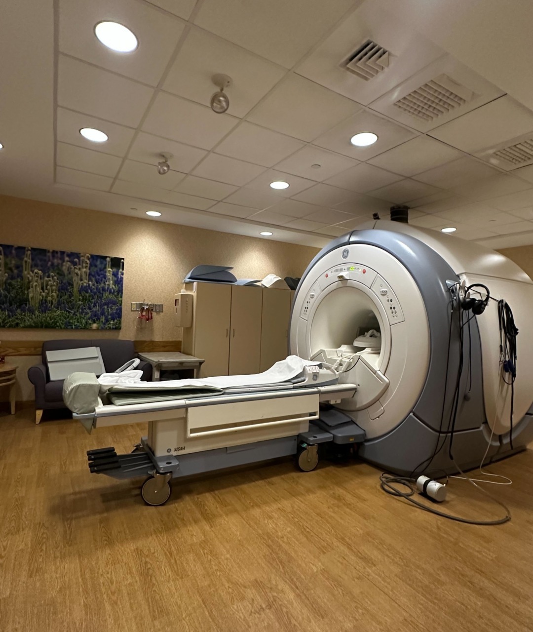 MRI machine in a hospital room with a patient bed nearby, overhead lights, and a calming wall mural in the background.
