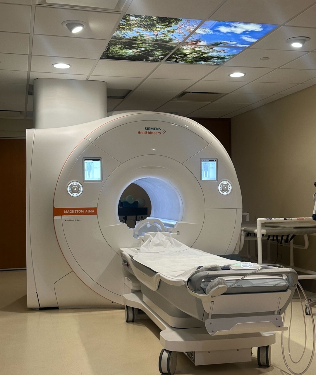MRI machine in a hospital room with a bed positioned for a scan, ceiling tiles display a serene nature scene.