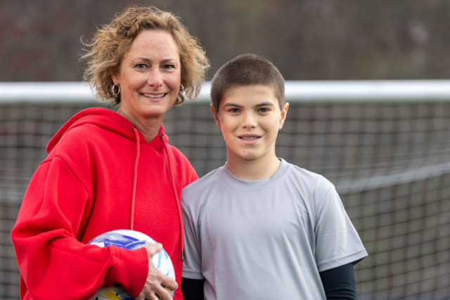 Woman in red hoodie holding a soccer ball, standing next to a young boy on a soccer field.