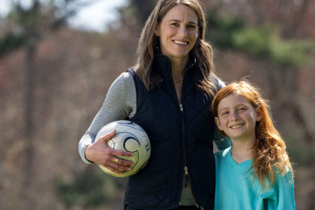 Woman holding a soccer ball stands outdoors with a smiling girl wearing a teal shirt.