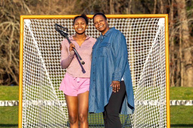 Two women smiling and standing in front of a lacrosse goal, one holding a lacrosse stick, outdoors.