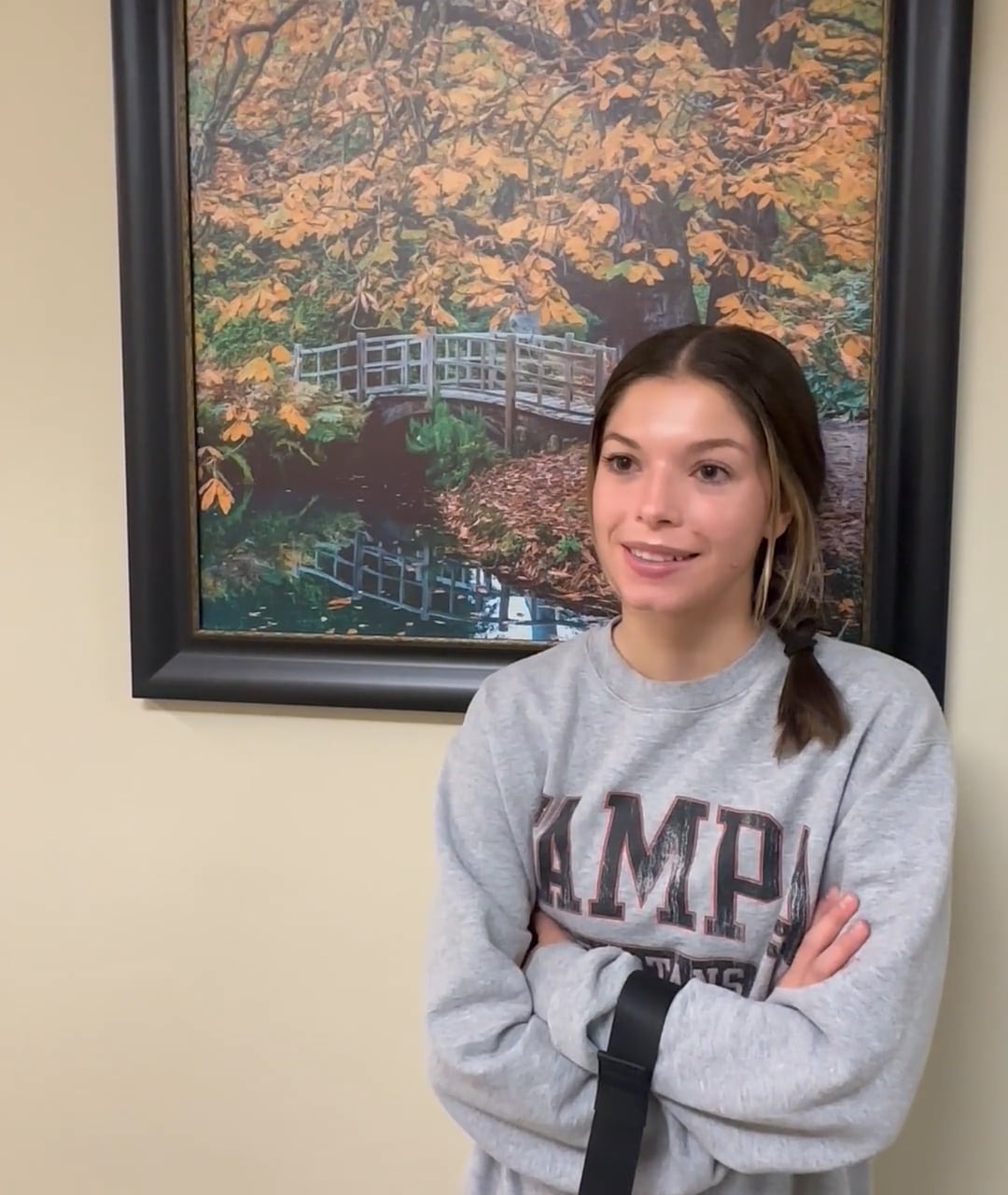 Person standing in front of a framed autumn landscape painting, wearing a casual gray sweatshirt indoors.